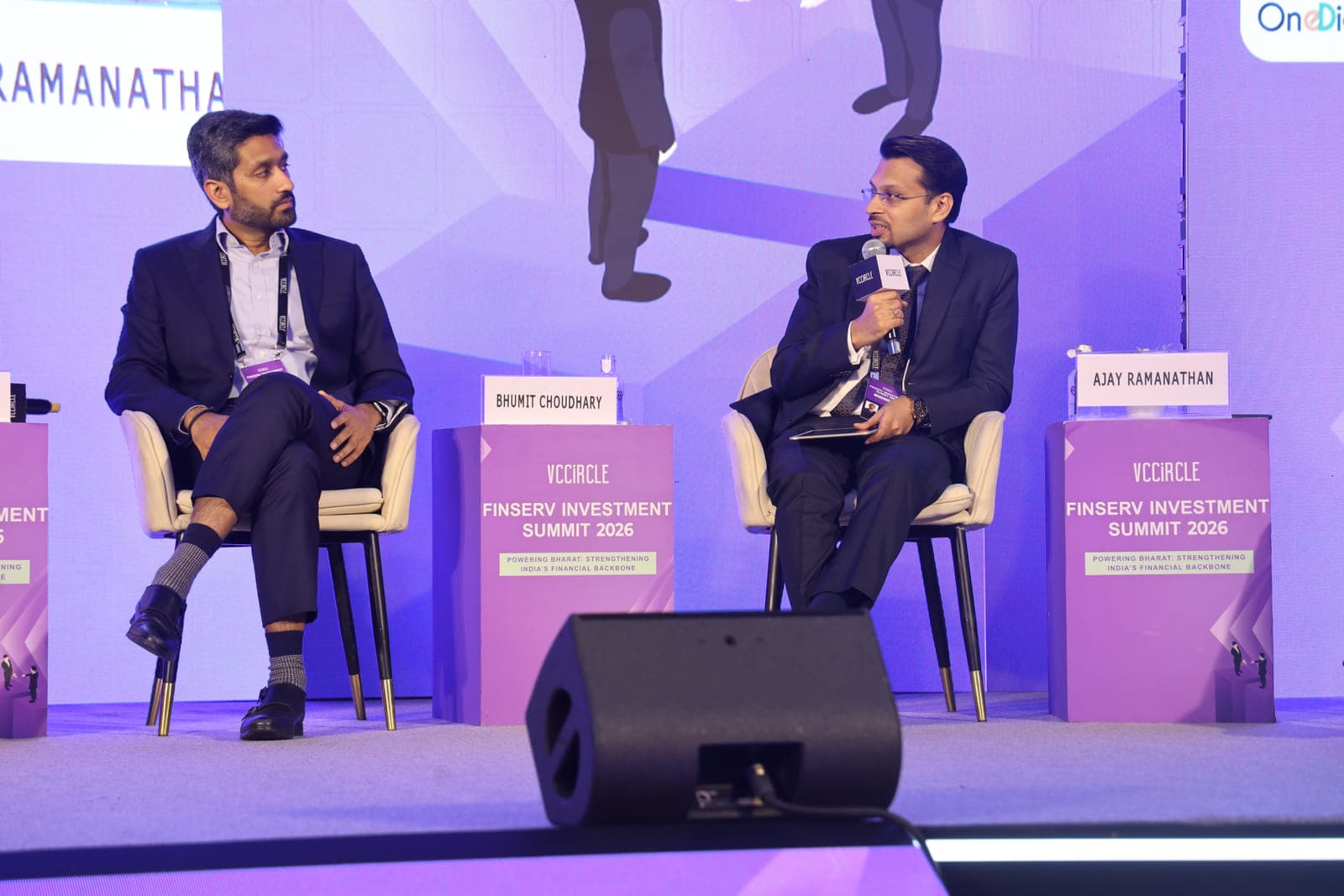 Two panellists seated on stage in front of a VCCircle backdrop, one speaking into a handheld microphone while the other listens.