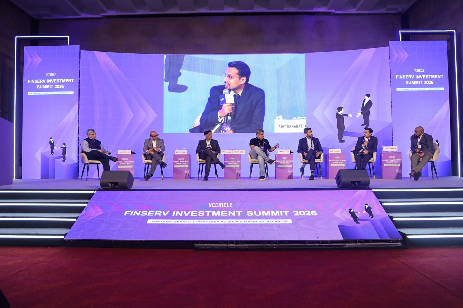 Seven speakers seated in a row across a conference stage, with the speaker in progress shown enlarged on the screen behind them.